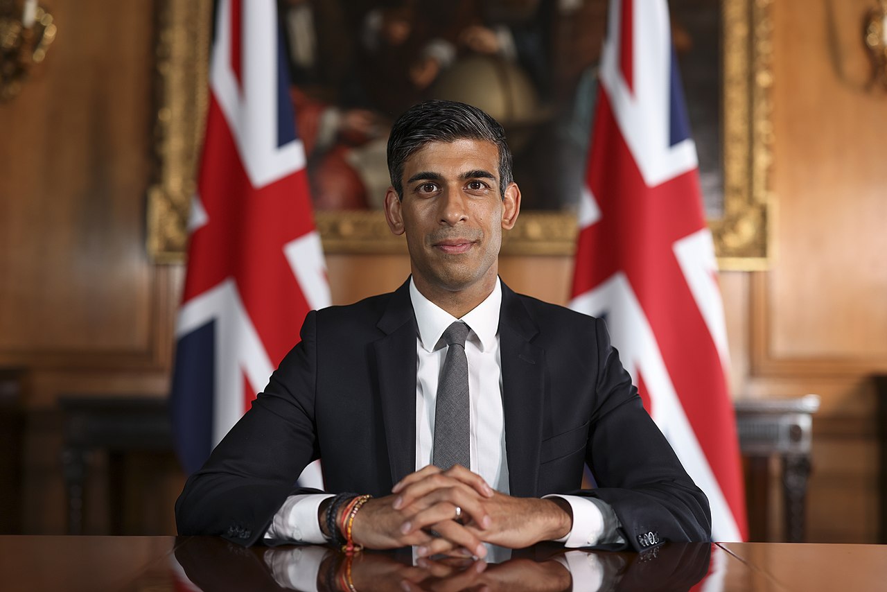 UK Prime Minister Rishi Sunak sitting at a desk, two UK flags in the background behind him.
